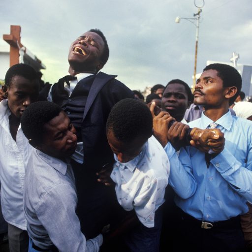 COPYRIGHT:  MAGGIE STEBER, PHOTOGRAPHER
MOTHER'S FUNERAL--A young Haitian man writhes in grief at the funeral for his mother in the National Cemetery in Pt-au-Prince, Haiti in November 1987.  His friends and family lend support in his moments of anguish.  His mother was killed in pre-election violence in Haiti in the weeks leading up to the first presidential elections in 30 years, following the fall of the cruel Duvalier regime in 1986.  All-night shooting could be heard in Haitian cities during the weeks leading up to the elections. On election day, polls opened and closed within a hour because of widespread slaughter of voters and the burning of ballots and polling stations.
