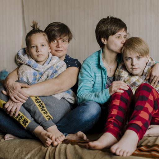 LVIV, UKRAINE - 21ST OF MARCH, 2022:  
Tamara Khrustalova (43, she/her, blue shirt) and Natalia Kravchenko (46, she/her, dark blue shit) live together and have two sons, Glib (12, red pants) and Oleksii (11, grey pants) and two cats Tima and Rysia. They sit on their bed in the flat of TamaraÕs grandmother on the 21st of March 2022. The  grandmother passed away a few years ago and now during the war they can use this home. They lived in Kyiv but fled to Lviv. When the air raid sirens go off they hide in the hallway. All the windows of the house are taped, so that when shelling or bombing starts, the glass does not fly into the home.
Tamara:
ÒWe travelled by train from Kyiv to Lviv, 2,5 weeks ago. We did not bring many belongings, we couldn't. We brought our clothes, our two cats and a computer. The train ride was really difficult, when we arrived to the train station there were hundreds of people waiting. They weren't selling tickets, they were evacuation trains and it was extremely busy. The lucky ones would get on, you really had to push your way through. It was so extremely crowded in the train, we needed to stand on one leg. The children found a small place in between seat to be seated and be a little more comfortable and safe. The train ride from Kyiv to Lviv took us around 11 hours. 
Now I am working at a LGBTQi refugee shelter, the shelter is set up by an organization called Insight. The people working in the shelter all know each other indirect from Kyiv. Our community is quite big, but many of us know each other. The shelter is specified for people from the LHBTQi community. I speak English, so that is quite helpful in this case. I clean the shelter, buy groceries or buy medicines. 
There are several quite specific medicines like different kind of hormones for instance for our trans community. Some of the medicines are donated from different countries like the Netherlands or other countries in Europe. Almost everyday I go to the drugstore to buy these medicines for the people in the shelter. 
Some of the medications for hormones are very specific, for instance the one that stops the growth of the larynx. These kind of medicines are not available here in Lviv, or not even in the neighboring countries like Poland. So as a volunteer, together with the team here I try to get this medicine from Germany. 
We also try to ship medicines and other things to the cities that are currently under siege. It is a difficult process, but these people need to be helped as well. One of the other things I do is help people cross the border from Ukraine to Poland. I have helped a family cross and in the near future I might be helping a trans person cross. There is someone staying at the LGBTQi shelter now that is trans. It is difficult for them to cross, because they have an 'M' in their passport, stating the fact they were born male. And males between the age of 18-60 are not allowed to leave the country. 
To be part of the LHBTQ community in Ukraine is really difficult. We don't really speak about being LGBTQi out in the open. It is more like a secret. Even our two sons, do not really know. I think they can guess, but we never told them anything. We are afraid they might tell other kids in school and that these kids will tell their parents. You just never know how people will react. And we do not want to put that burden on our children.
We are not openly affectionate on the streets together, we can hold hands, because people might think we are friends. But we canÕt kiss in the street. We do feel like a normal family, with a normal life and that is also how we behave,. Except the fact we won't be openly affectionate outside. This is just not part of the possibilities within this country at the moment. 
We have been together for 15 years and we believe the rights of LGBTQi's are very important, but we do not demonstrate, or go to pride. I don't want to get into problems, I have a family. There are people who come to the march just to hurt LGBTQi's. There is a lot of police to prevent this, but it still happens. I don't want to be there. I wish Ukraine was more tolerant towards the LGBTQi community. But this is one of the many problems our country faces. The war does not change this for me, but what I do feel is happening during the war is the fact that LGBTQi's are sticking out for each other and others. The community is really there for each other, and stick together. That is beautiful.Ó

Many LGBTQI's left their hometowns during the invasion  of Russia into Ukraine. Just like other Ukrainians they are fleeing the attacks. 
The LGBTQI- community is said to be targets of a Russian oppression campaign and names of LGBTQI-activists have apparently appeared on kill-lists. While fleeing, many of them made sure to stay close together as a community, to feel the safety of being with like-minded people. Some of them set up shelters themselves (including the distributing HIV and hormone medication) and volunteer there. 
The trans-people have an added issue when it comes to the war. In many cases trans-women have a 'M' in their passport, stating they were born 'male'. Even though they might have undergone hormone-therapy in recent years and might feel (and be) female all the way; on paper they aren't. These trans-women cannot leave the country, as they are seen as 'men' by the Ukrainian officials, who will not let them cross the border. Men between the age of 18-60 have to stay and preferable join the army. 
Russia invaded Ukraine on the 24th of February 2022.