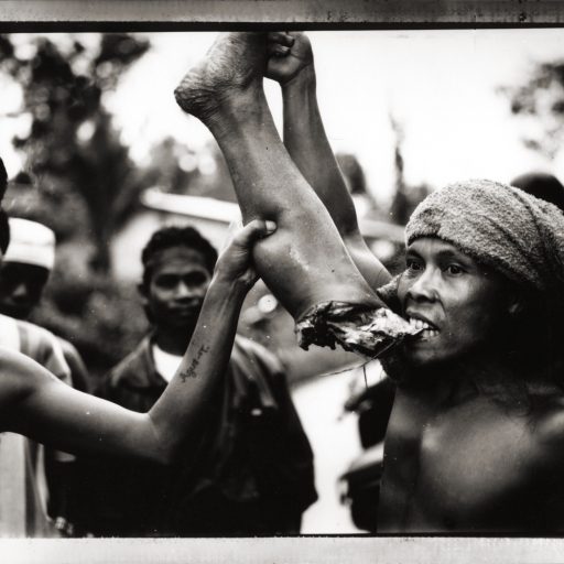 A Dayak warrior gnaws on the leg of a slain Madurese settler, near Singkawang, Kalimantan, Indonesian Borneo. March 1999