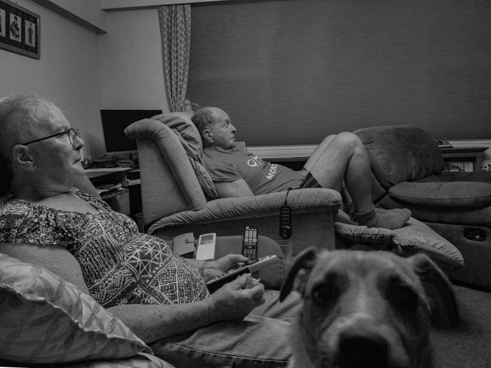 Beverley and Andrew Davy (husband and wife) relax watching television as Maggie the dog spots the camera. 4th February 2022, Auroa, Taranaki, Aotearoa. Photo by Nick Netzler