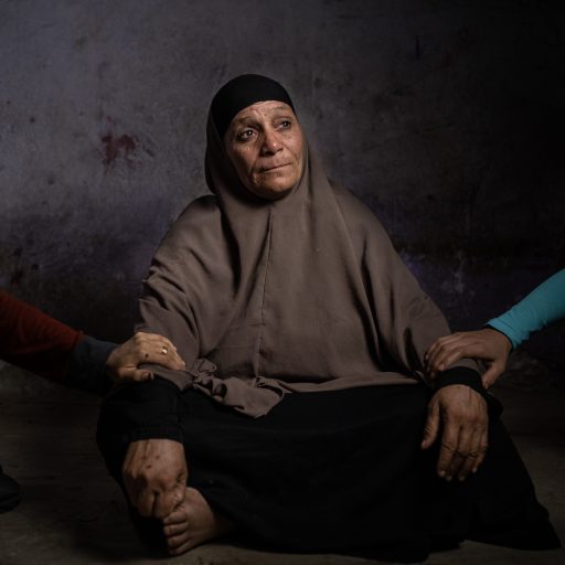 A woman Ajab Muhammad Miftah, 54, poses for a portrait in his house where she lives, in the village of  Ezbat Soliman, Fayoum, Egypt, Sep 22, 2022. Two hands of her daughter support her after she lost her husband and two of his young sons in a microbus accident during their trip to Aswan, The trips of the fishermen from Fayoum governorate, in search of a living, in Lake Nasser in the Aswan governorate, which is more than 825 km from Fayoum, turned into trips for death after they had to abandon Lake Qarun, from which they used to fish, due to the high pollution rates in the area. ©Fatma Fahmy for the VII Mentor Program.