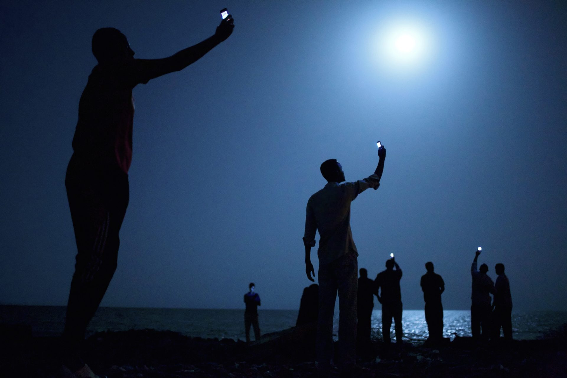 Impoverished African migrants crowd the night shore of Djibouti city on February 26, 2013, trying to capture inexpensive cell signals from neighboring Somalia—a tenuous link to relatives abroad. ©John Stanmeyer / VII.