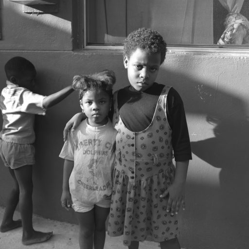 Overtown, Miami, 1992.
Portrait of sisters outside their apartment in a housing complex in one of The City’s oldest Black communities. Called “Colored Town” in The Jim Crow South because it was the only place Black residents were allowed to live. Overtown became the heart of Miami’s Black Community, with thriving Black-owned businesses. When great Black artists like Billie Holiday and Nat King Cole traveled to perform in clubs on Miami Beach, the Beach hotels were segregated, and these legendary performers traveled across the causeway to stay in Overtown. The housing and municipal infrastructure has traditionally remained marginalized, and in the 1960s, urban renewal literally put a freeway through Overtown, thus physically passing over the neighborhood and perpetuating layers of segregation that remain today. © Brenda Ann Kenneally.
