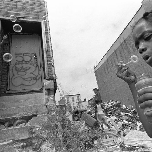 Bushwick, Brooklyn, 1999.
Children play in one of the many abandoned empty lots on Dodworth Street. Gentrification began to sweep the neighborhood in 2006, and the house in the background has been rebuilt and sold as triple-stack condos for over one million dollars per unit. All of the residents in this neighborhood have been priced out, and essentially, the community as we knew it has vanished. © Brenda Ann Kenneally.