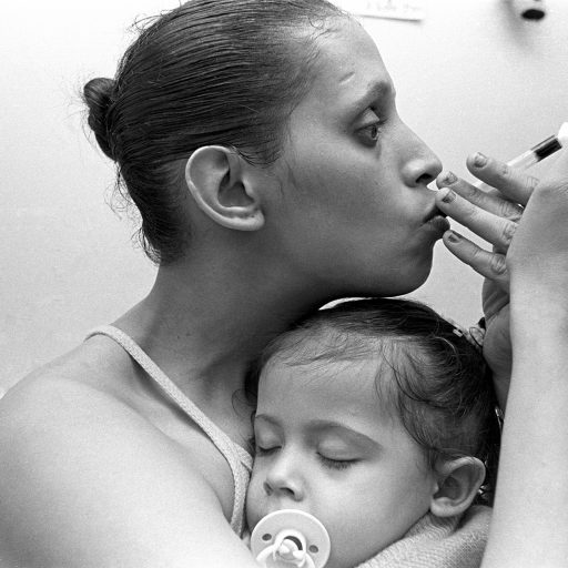 Bushwick, Brooklyn, 2000.
A crack-addicted mother sneaks a hit from her stem while her toddler daughter naps in her arms.
Pre-gentrified Bushwick had become one of the poorest neighborhoods in Brooklyn and ripe for the crack era addiction and desperation that intersected with other problems in the low-status neighborhood. By 2010, Bushwick had become one of the most expensive places to live in the USA and internationally synonymous with all things hip and trendy. None of the community households from the 1990s remain on the Dodworth Street block where this picture was made. © Brenda Ann Kenneally.