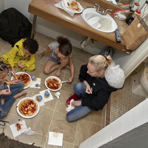 Menands, New York, 2020.
A mother feeds her children on the bathroom floor in their room at The Schuyler Inn Homeless Shelter. During the first month of Covid lockdown, there were no masks or places for social distancing in congregate facilities like shelters. The Inn closed its dining room, and there were no tables in rooms, as food privileges in individual quarters had been suspended years earlier to prevent the constant infestations of roaches. The bathroom floor seemed to be the only place with a surface that could be sanitized. © Brenda Ann Kenneally.
