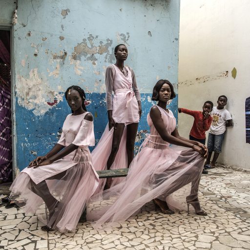 Curious residents and a street vendor selling material look on as models (L-R) Diarra Ndiaye, Ndeye Fatou Mbaye and Malezi Sakho wear outfits by Senegalese designer Adama Paris in the Medina neighbourhood of Senegal's capital, Dakar, December 15, 2018.  © Finbarr O’Reilly.