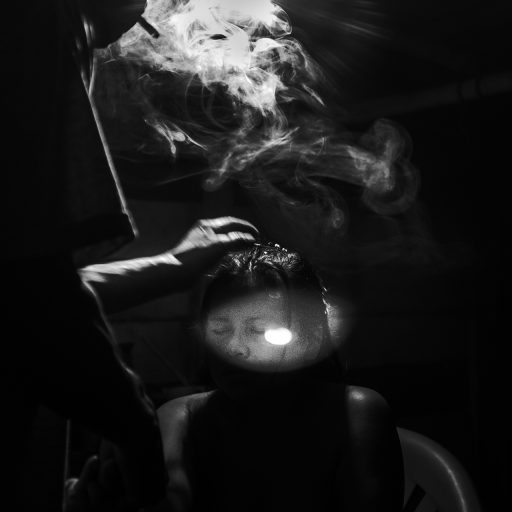 In May, 2017, Aladino, a native Bora shaman, blows smoke on his patient's head during one of the healing sessions late at night. The house is located in a small village in the Amazon. Before blowing the smoke, he performs a prayer in Bora language and asks the spirits to guide him throughout the process. The spiritual process is to make a woman called Marili, who feels sick from her stomach and is facing trouble sleeping, better again. Aladino says that the duty of a natural healer is to work with all kind of diseases from headaches, which can take a day, to the worse like malediction, which can take weeks. © Leonardo Carrato.