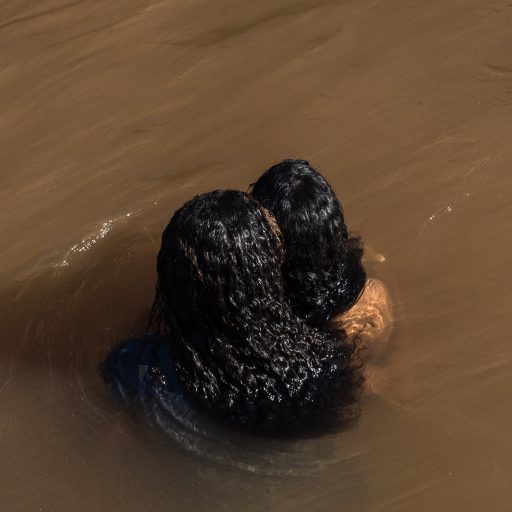 On November 17, 2020, Larissa, 15 years old, and Isabela Pataxó, 16 years old, embrace in a stream in the village where the former indigenous prison, the Krenak Reformatory, was located in the city of Carmésia, state of Minas Gerais, Brazil. Water and the passage of a river are sacred according to indigenous culture, the lifeblood of the community. This small stream, the only source of water in the village, does not provide the essence necessary for the survival of the community, and vital activities such as fishing are not possible. This feeling of not belonging affects the psychology of the new generation that is on a quest to regain their indigenous identity. Faced with an oppressive past and an uncertain present, staying together through physical closeness is one of the few options to build a sense of community. © Leonardo Carrato.