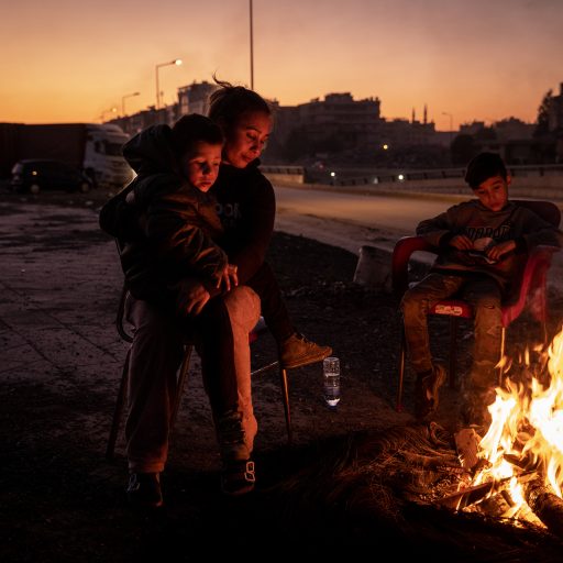 Gulbahar Bicer, 35, holds her son Deniz, 5, while her other son Ali, 10, tends watches fire outside their tent shelter in Iskenderun, Turkey, on Thursday, February 16, 2023. The single mother of the children, Gulbahar, lost her home in the major earthquakes which hit Turkey and Syria on February 6. Ms. Bicer says that her application for a tent from a Turkish organization is still being processed. In the mean time, she is caring for her three children and mother-in-law by herself, living in a tent on the side of a main road in Iskenderun. Ms. Bicer says her son Deniz is experiencing panic attacks after the earthquake, and keeping her children clean without running water or electricity is the biggest challenge. © Nicole Tung.