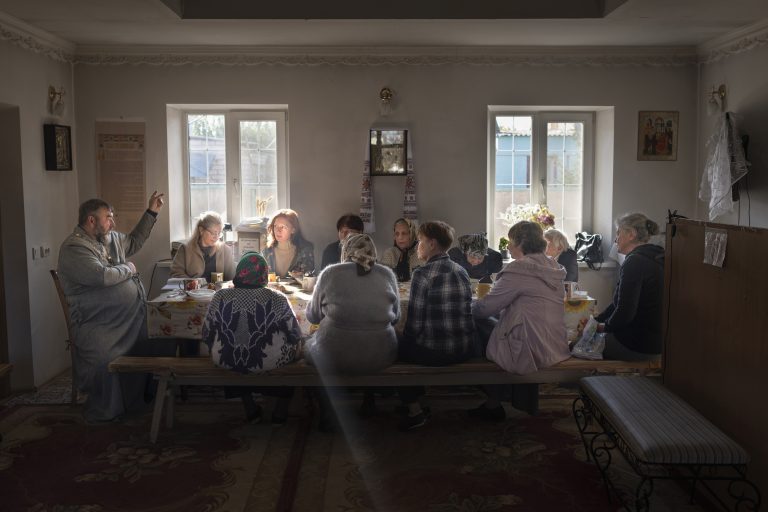 Parishioners gather around a table in a small chapel after morning prayers. Every week, residents come together in this chapel for the Sunday sermon following the flooding of their church due to the Kakhovka Dam breach on Jun. 6, 2023. The flood waters produced by the breach submerged numerous towns and villages in Kherson Oblast. Many international experts and Ukrainian officials say the evidence suggests Russian forces, which captured the dam in the early days of the invasion and remained in control at the time of the disaster, were responsible, a claim that Russia denies. Nov. 5, 2023, Kherson, Ukraine. © Oleksandr Rupeta / VII.