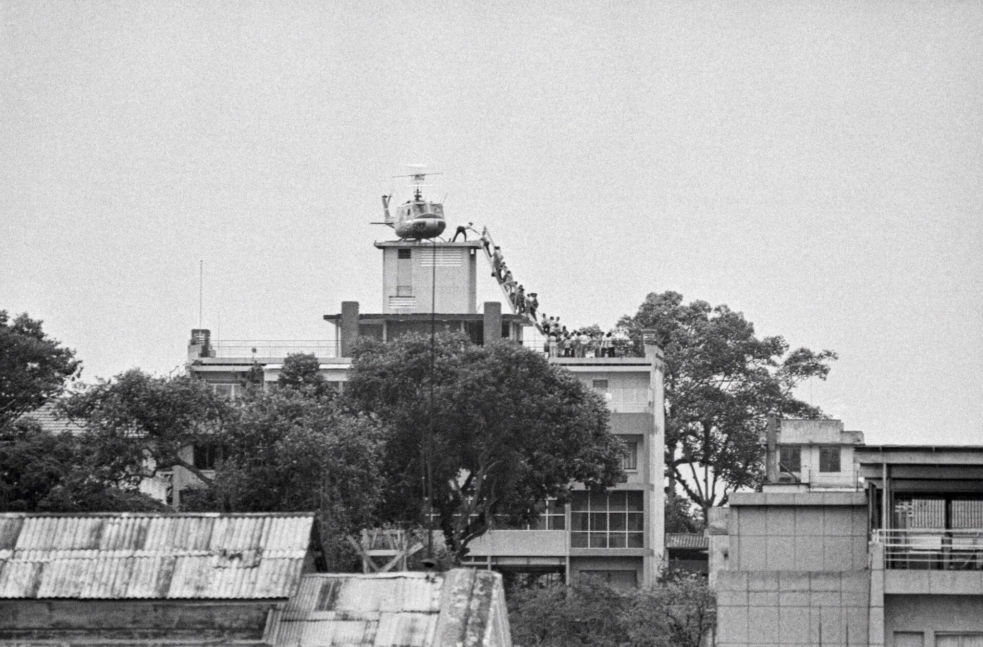 In what has become a defining image of the fall of Saigon, a man believed to be a CIA employee helps Vietnamese evacuees onto a helicopter from the top of 22 Gia Long Street, an apartment building a half mile from the U.S. Embassy. The helicopter was operated by Air America, an airline covertly owned and operated by the CIA from 1950-1976. Air America planes were masqueraded as civilian aircraft when, in actual fact, they were supporting U.S. Special Forces in secret operations across Asia. They delivered weapons and military equipment to American allies in Laos, Cambodia, Vietnam, and flew covert missions as far as Burma and China. When the image was first published, it was often incorrectly assumed to show Americans being evacuated from the American Embassy in the final hours of the war. / Dans ce qui est devenu une image phare de la chute de Saigon, un homme soupçonné d'être un employé de la CIA aide les Vietnamiens évacués à monter à bord d'un hélicoptère depuis le 22, rue Gia Long, un immeuble situé à 800 mètres de l'ambassade américaine. L'hélicoptère était exploité par Air America, une compagnie aérienne secrètement détenue et exploitée par la CIA de 1950 à 1976. Les avions d’Air America étaient déguisés en avions civils alors qu’en réalité, ils soutenaient les forces spéciales américaines dans des opérations secrètes à travers l’Asie. Ils ont livré des armes et du matériel militaire aux alliés américains au Laos, au Cambodge, au Vietnam, et ont effectué des missions secrètes jusqu’en Birmanie et en Chine. Lorsque l’image a été publiée pour la première fois, on a souvent supposé à tort qu’elle montrait des Américains évacués de l’ambassade américaine dans les dernières heures de la guerre. Photo by Hugh Van Es/Bettmann Archive via Getty Images.