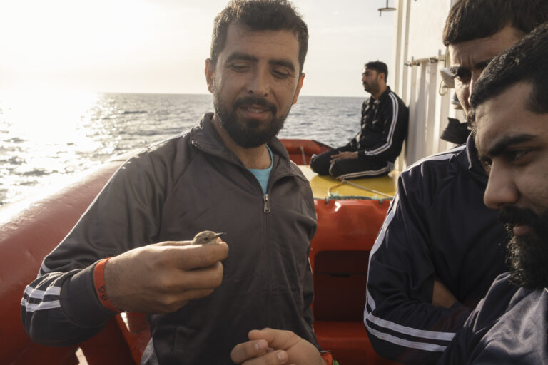 A rescued man holds a small bird he found drifting at sea before being saved by the Ocean Viking. Survivors often spend days exposed to the elements in fragile wooden boats before help arrives. Following rescues, Italian authorities frequently assign distant ports of disembarkation—requiring up to five days of navigation—delaying medical care and draining the ship’s resources, despite the often critical physical and psychological condition of those on board. © Javier Alvarez.