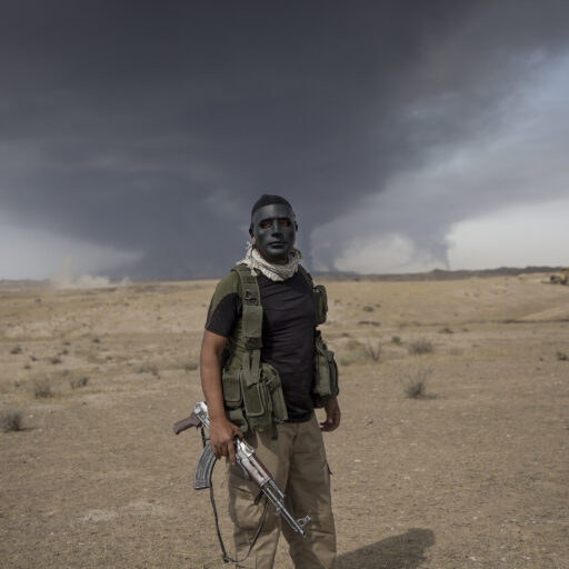 A portrait of an Iraqi resistance fighter in Kirkuk, Iraq, with black smoke rising behind him from oil wells blown up by ISIS. © Ali Khara.