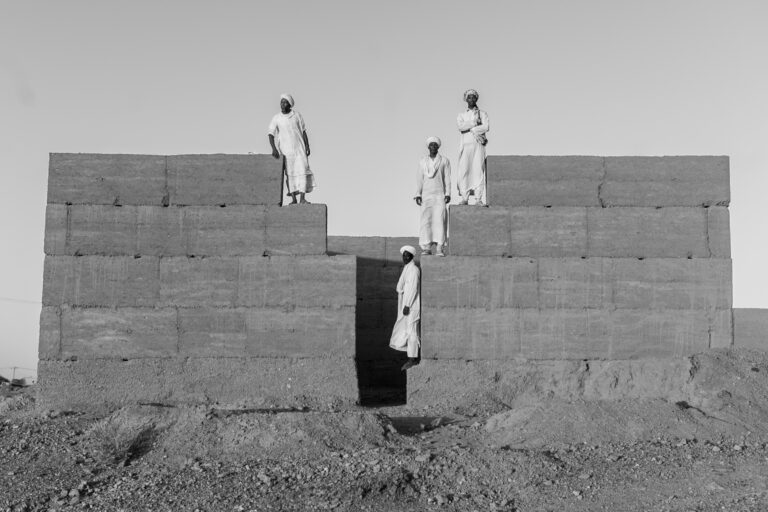 Young Gnawa men stand in their village of Khamlia, a community shaped by centuries of trans-Saharan movement. Descendants of people brought north across the desert through forced migration and exile, they live with histories of movement that continue to shape identity, belonging, and daily life today. Khamlia, Moroccan Sahara, Morocco, 2025. © Maroussia Mbaye.